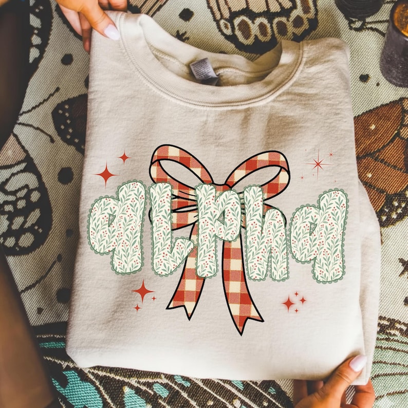 Pode incluir: Uma camisola dobrada, de cor creme, com a palavra "ALPHA" em uma fonte decorativa. As letras s&atilde;o preenchidas com um padr&atilde;o floral verde e vermelho e delineadas com uma borda ondulada. Um la&ccedil;o xadrez vermelho e branco est&aacute; centrado acima da palavra, com estrelas vermelhas.