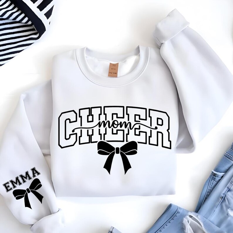 May include: White sweatshirt with the text "CHEER mom" in black block letters and a black bow. The left sleeve has the name "EMMA" and a black bow. The sweatshirt is folded on a white surface.