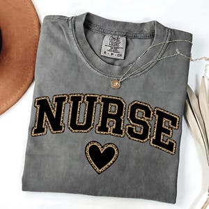 May include: Gray Comfort Colors t-shirt with the word "NURSE" in black block letters with a leopard print fill. A leopard print heart is below the word. A gold necklace with a small pendant is on the shirt.