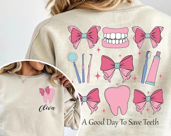 May include: Beige sweatshirt with a dental theme. The design includes pink bows, teeth, dental tools, a toothbrush, and toothpaste. The text "A Good Day To Save Teeth" is printed on the bottom. A smaller image on the front has a tooth with a bow and the name "Oliva".