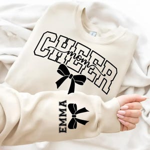 May include: Cream-colored sweatshirt with the words "CHEER mom" in black block letters and a black bow graphic. The sleeve displays the name "EMMA" and a matching bow. The sweatshirt is folded on a white surface.