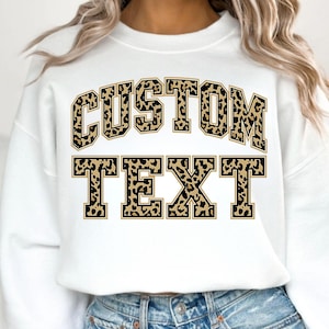 May include: White sweatshirt with the words "CUSTOM TEXT" in a leopard print design. The text is in a bold, collegiate-style font, with a black outline. The sweatshirt is a casual, everyday garment.