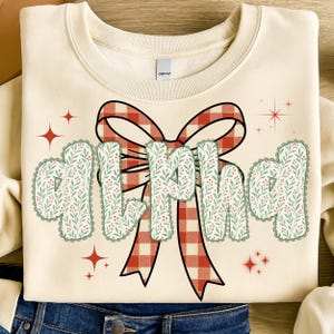 Pode incluir: Camisola creme com a palavra "GIRL MAMA" em uma fonte decorativa. As letras t&ecirc;m uma borda ondulada e s&atilde;o preenchidas com um padr&atilde;o floral verde e vermelho. Uma grande fita xadrez vermelha e branca est&aacute; atr&aacute;s do texto, com detalhes de estrelas vermelhas.