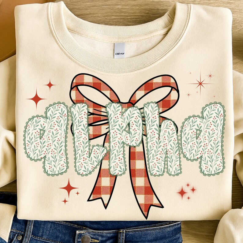 Pode incluir: Camisola creme com a palavra "GIRL MAMA" em uma fonte decorativa. As letras t&ecirc;m uma borda ondulada e s&atilde;o preenchidas com um padr&atilde;o floral verde e vermelho. Uma grande fita xadrez vermelha e branca est&aacute; atr&aacute;s do texto, com detalhes de estrelas vermelhas.