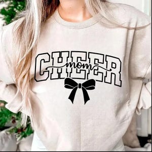 May include: A beige sweatshirt with the word "CHEER" in a block letter style, with "mom" in a cursive font in the middle. A black bow is below the word "mom".