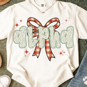 Pode incluir: Camiseta creme com a palavra "MILF" em uma fonte decorativa. As letras s&atilde;o adornadas com um padr&atilde;o floral verde e branco e contornadas com uma borda ondulada. Uma fita xadrez vermelha e branca est&aacute; atr&aacute;s da palavra.
