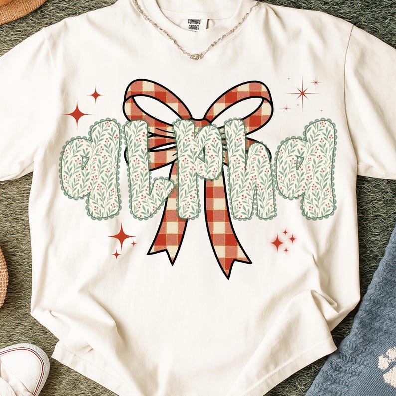 Pode incluir: Camiseta creme com a palavra "MILF" em uma fonte decorativa. As letras s&atilde;o adornadas com um padr&atilde;o floral verde e branco e contornadas com uma borda ondulada. Uma fita xadrez vermelha e branca est&aacute; atr&aacute;s da palavra.