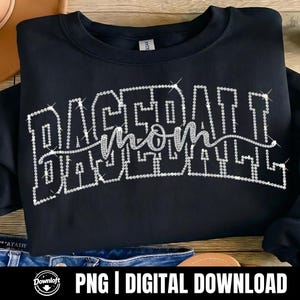 May include: Black sweatshirt with the text "BASEBALL MOM" in a sparkling, rhinestone-like design. The sweatshirt is folded, and the image is set against a wooden background. The text is in a script font.