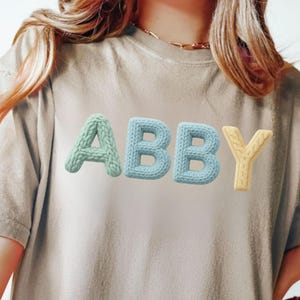 Op de afbeelding: Een beige t-shirt met de naam "ABBY" in 3D gebreide letters. De letters zijn in pastelkleuren: mintgroen, lichtblauw en geel. Het shirt heeft korte mouwen en een ronde hals.