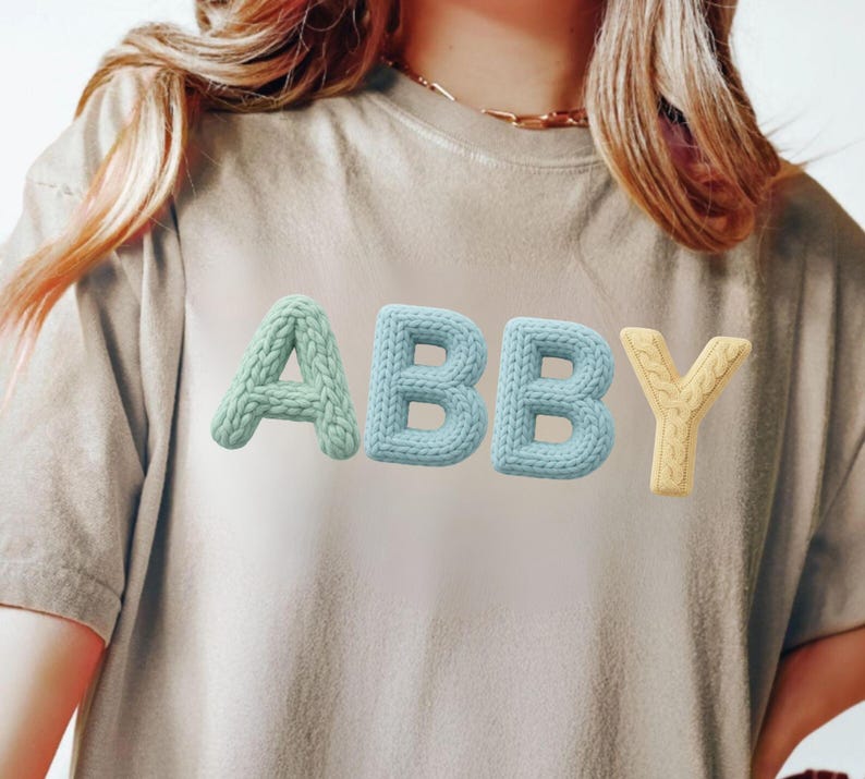 Op de afbeelding: Een beige t-shirt met de naam "ABBY" in 3D gebreide letters. De letters zijn in pastelkleuren: mintgroen, lichtblauw en geel. Het shirt heeft korte mouwen en een ronde hals.