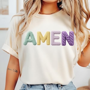 Op de afbeelding: Cr&egrave;mekleurig T-shirt met het woord "AMEN" in kleurrijke, getextureerde letters. De letters zijn in pastelkleuren: lichtgroen, geel, lichtpaars en paars. De letters lijken gebreid of gehaakt.