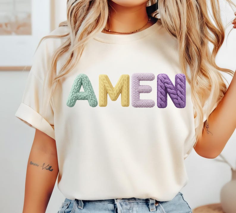 Op de afbeelding: Cr&egrave;mekleurig T-shirt met het woord "AMEN" in kleurrijke, getextureerde letters. De letters zijn in pastelkleuren: lichtgroen, geel, lichtpaars en paars. De letters lijken gebreid of gehaakt.
