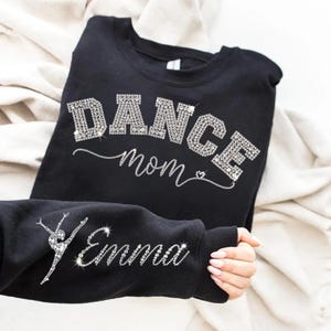 Könnte beinhalten: Schwarzes Sweatshirt mit dem Text "DANCE mom" in Strassbuchstaben. Der Ärmel zeigt eine Strass-Tänzerin und den Namen "Emma". Das Sweatshirt ist auf einer weißen Oberfläche gefaltet.
