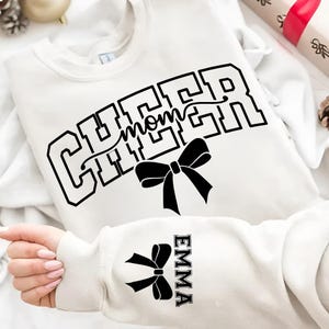 Puede incluir: Sudadera blanca con las palabras "CHEER mom" en letras negras y un lazo negro. La manga presenta un lazo a juego y el nombre "EMMA" en fuente vertical. Es una prenda de vestir informal.