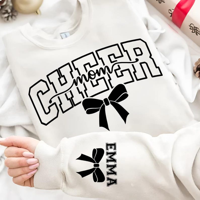 May include: A white sweatshirt with the words "CHEER mom" in black block letters and a black bow graphic. The sleeve features a matching bow and the name "EMMA" in a vertical block font. The sweatshirt is a casual apparel item.