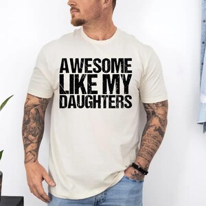 Op de afbeelding: Cr&egrave;mekleurig T-shirt met de tekst "AWESOME LIKE MY DAUGHTERS" in vette, zwarte letters. Het shirt heeft korte mouwen en een ronde hals. De persoon die het shirt draagt heeft tatoeages op zijn armen en draagt een blauwe jeans.