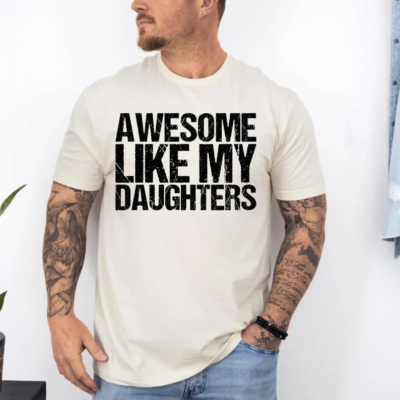 Op de afbeelding: Cr&egrave;mekleurig T-shirt met de tekst "AWESOME LIKE MY DAUGHTERS" in vette, zwarte letters. Het shirt heeft korte mouwen en een ronde hals. De persoon die het shirt draagt heeft tatoeages op zijn armen en draagt een blauwe jeans.