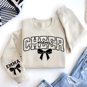 May include: Cream-colored sweatshirt with the words "CHEER MOM" in black block letters and a black bow graphic. The sleeve features the name "EMMA" and a matching bow. The sweatshirt is paired with blue jeans.