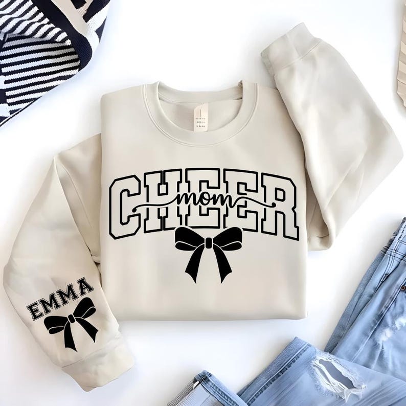 May include: Cream-colored sweatshirt with the words "CHEER MOM" in black block letters and a black bow graphic. The sleeve features the name "EMMA" and a matching bow. The sweatshirt is paired with blue jeans.