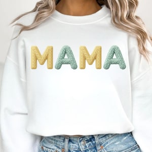 Op de afbeelding: Witte sweatshirt met het woord "MAMA" in getextureerde, 3D-letters. De letters "M" en "A" zijn geel, terwijl de middelste "A" lichtblauw is. De sweatshirt heeft een casual stijl.