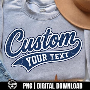 May include: A light gray crewneck sweatshirt featuring a large, dark navy blue graphic with a distressed texture. The design displays the word Custom in a bold, cursive script font, with the words YOUR TEXT printed in a block font below.