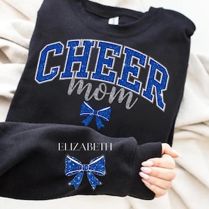 Peut inclure: Sweat-shirt noir avec l'inscription "CHEER MOM" en paillettes bleues, le mot "mom" en paillettes argentées. Un nœud bleu à paillettes est en dessous. La manche porte le nom "ELIZABETH" et un nœud bleu à paillettes.