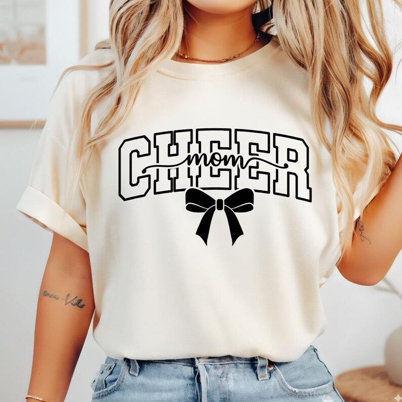 May include: Cream-colored t-shirt with the words "CHEER mom" in black block letters and a black bow graphic. The shirt has short sleeves and a relaxed fit.