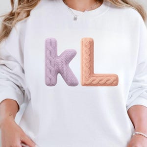 Op de afbeelding: Wit sweatshirt met de letters "KL" in een 3D kabelgebreid ontwerp. De letter "K" is zacht lavendelkleurig, terwijl de letter "L" perzikkleurig is. De letters staan in het midden op de voorkant van het sweatshirt.