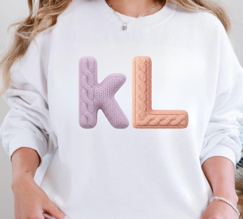 Op de afbeelding: Wit sweatshirt met de letters "KL" in een 3D kabelgebreid ontwerp. De letter "K" is zacht lavendelkleurig, terwijl de letter "L" perzikkleurig is. De letters staan in het midden op de voorkant van het sweatshirt.