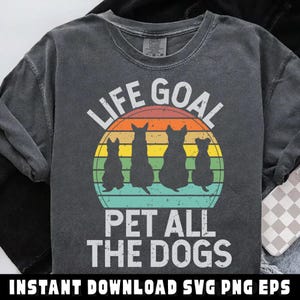 May include: Dark gray t-shirt with the text "LIFE GOAL PET ALL THE DOGS" in white. The design features four black dog silhouettes against a retro sunset background. The shirt is a comfortable, casual style.