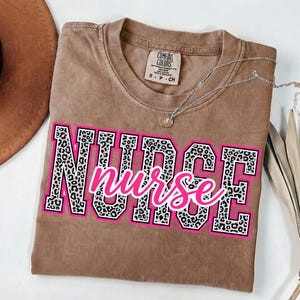 May include: A brown Comfort Colors t-shirt with the word "NURSE" in large, leopard print letters outlined in pink and white. The word "nurse" is written in cursive in pink over the top. A silver necklace with a small pendant is draped across the shirt.