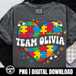 以下が含まれることがあります： カラフルなパズルハートのデザインと「TEAM OLIVIA」の文字が入った濃いグレーのTシャツ。ハートは虹、手形、花で囲まれています。シャツの下部には「PNG | DIGITAL DOWNLOAD」と書かれています。