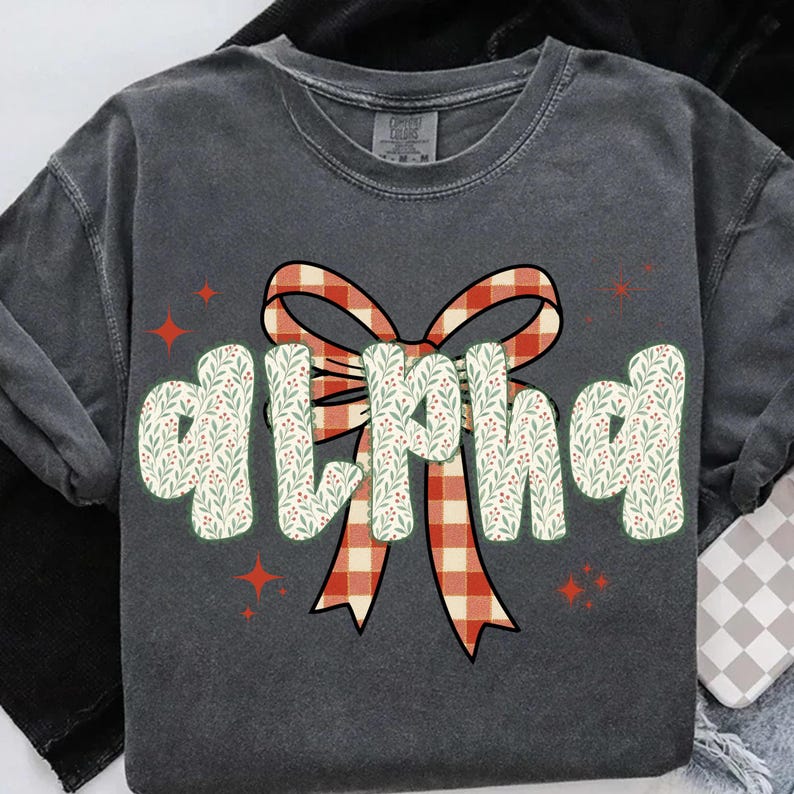 Pode incluir: Camiseta cinza escuro de manga comprida com a palavra "ALPHA" em uma fonte extravagante. As letras s&atilde;o decoradas com um padr&atilde;o floral branco e verde. Uma fita xadrez vermelha e branca est&aacute; atr&aacute;s da palavra, com detalhes de estrelas vermelhas.