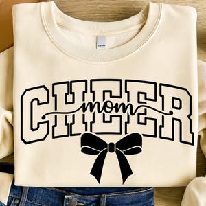 May include: Cream-colored sweatshirt with the word "CHEER mom" in black block letters and a black bow graphic. The sweatshirt is folded, with the bottom hem visible, resting on a denim surface.