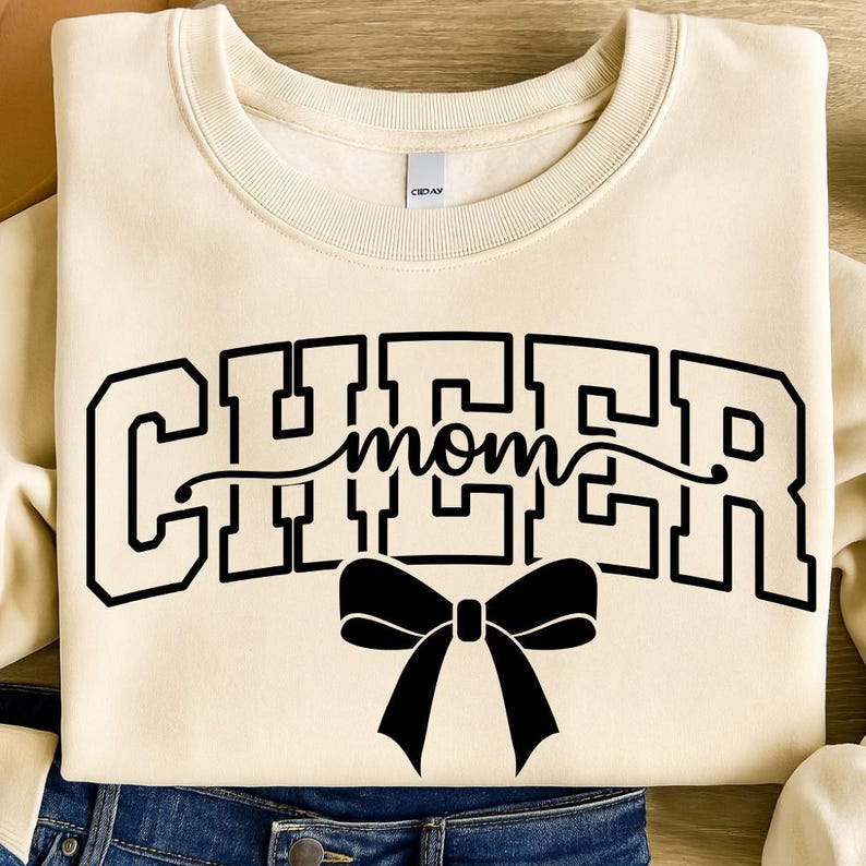 May include: Cream-colored sweatshirt with the word "CHEER mom" in black block letters and a black bow graphic. The sweatshirt is folded, with the bottom hem visible, resting on a denim surface.