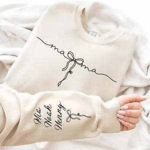 May include: Cream-colored sweatshirt with the word "mama" in cursive, connected by a bow graphic. The sleeve displays the names "Mia", "Noah", and "Henry" in cursive, with a bow graphic. The sweatshirt is folded on a white surface.