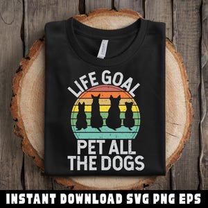 May include: Black t-shirt with the text "LIFE GOAL PET ALL THE DOGS" in white. The design features four dog silhouettes in front of a retro sunset. The shirt is folded on a wooden surface.