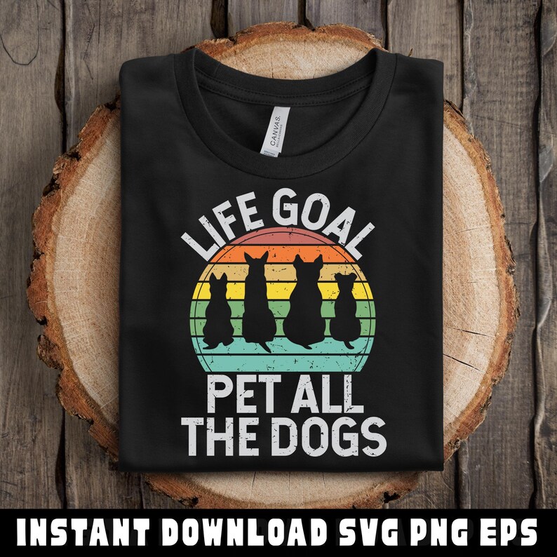 May include: Black t-shirt with the text "LIFE GOAL PET ALL THE DOGS" in white. The design features four dog silhouettes in front of a retro sunset. The shirt is folded on a wooden surface.