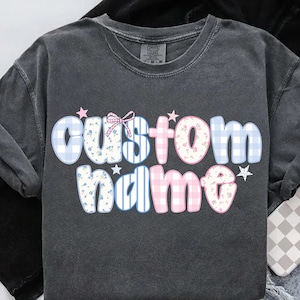 Puede incluir: Sudadera de manga larga gris oscuro con la frase "custom name". Las letras están adornadas con estampados florales, cuadros y rayas en tonos azules, rosas y blancos, con detalles de estrellas. Un lazo está encima de la palabra "custom".