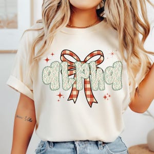 Pode incluir: Camiseta creme com a palavra "ALPHA" em uma fonte decorativa e um la&ccedil;o xadrez. As letras s&atilde;o contornadas com um design floral e o la&ccedil;o &eacute; vermelho e branco. Acentos de estrelas vermelhas est&atilde;o espalhados ao redor do texto.