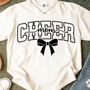 May include: Cream-colored t-shirt with the word "CHEER" in bold, black, block letters, with "mom" in cursive script in the middle. A black bow is centered below the text. The shirt has a crew neck.