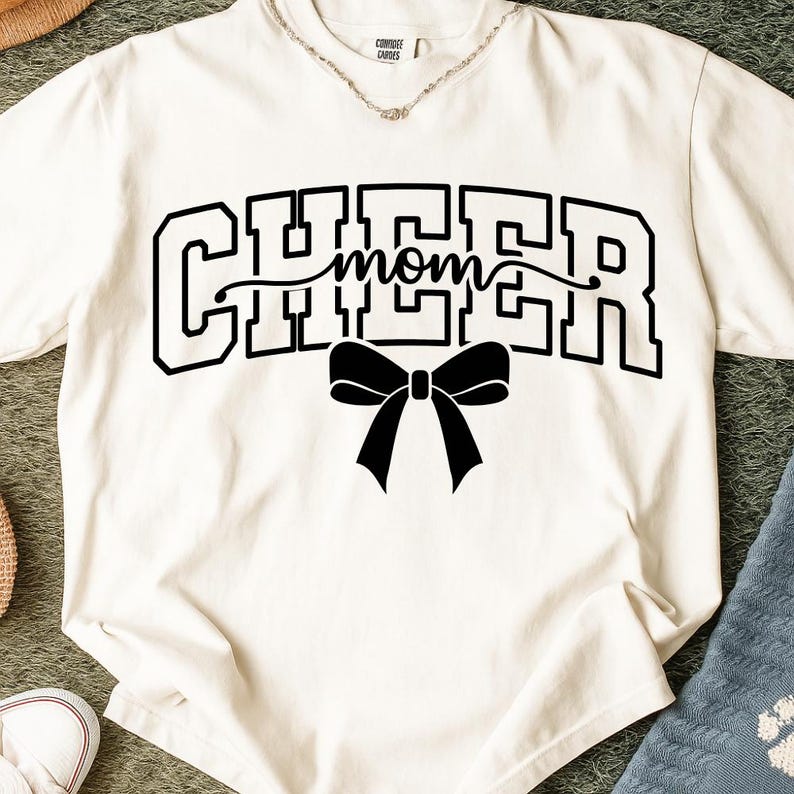 May include: Cream-colored t-shirt with the word "CHEER" in bold, black, block letters, with "mom" in cursive script in the middle. A black bow is centered below the text. The shirt has a crew neck.