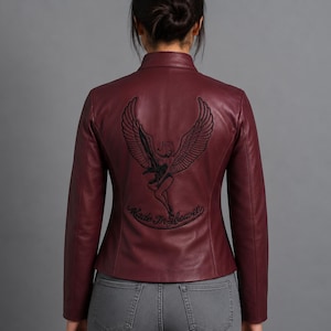 Peut inclure: Une veste bordeaux à col officier et manches longues. Le dos présente une grande broderie noire représentant une silhouette ailée avec l'inscription Made In Heaven en arc de cercle. La veste est portée sur un jean en denim gris.