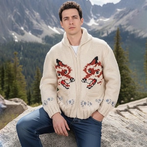 May include: Cream-coloured zip-up cardigan sweater with a fox design in red and black on the front. The sweater has a shawl collar, two pockets, and blue and grey accents on the cuffs and hem. The person is wearing blue jeans.
