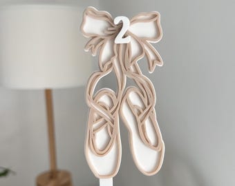 Op de afbeelding: Een gelaagde, gebroken witte en bruine ballet schoen decoratie met een strik en het nummer 2. Het ontwerp toont twee balletschoenen die aan een strik hangen. Het nummer 2 staat in het midden van de strik.