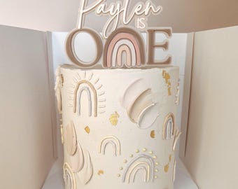 May include: A white frosted cake decorated with pastel rainbows and gold accents. The cake is topped with a custom cake topper that reads "Payten is One". The cake has a smooth finish with textured buttercream details.