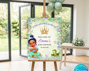 May include: A rectangular baby shower welcome sign on a wooden easel featuring a gold crown, a cartoon character in a green dress, and a green frog. The text reads: A little princess is on the way, WELCOME TO Emma's BABY SHOWER, 07.19.2026.