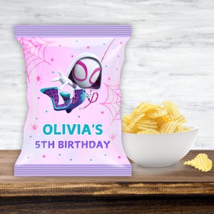 May include: A pink party favor bag featuring a Spider-Gwen character illustration, spiderwebs, and the text "OLIVIA'S 5TH BIRTHDAY." A white bowl holds a serving of ruffled potato chips, with one chip resting on the wooden surface.