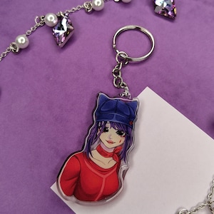 May include: An acrylic keyring featuring an anime-style character with purple hair, wearing a blue cap and a red long-sleeved shirt with a red neck scarf. The character has a small red mark on their cheek. The keyring includes a silver metal ring.