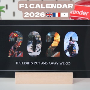 Peut inclure: Calendrier F1 noir pour 2026, avec des images à l'intérieur des chiffres. Le calendrier a une base en bois et une bordure à damier. Le texte sur le calendrier se lit "IT'S LIGHTS OUT AND AWAY WE GO."